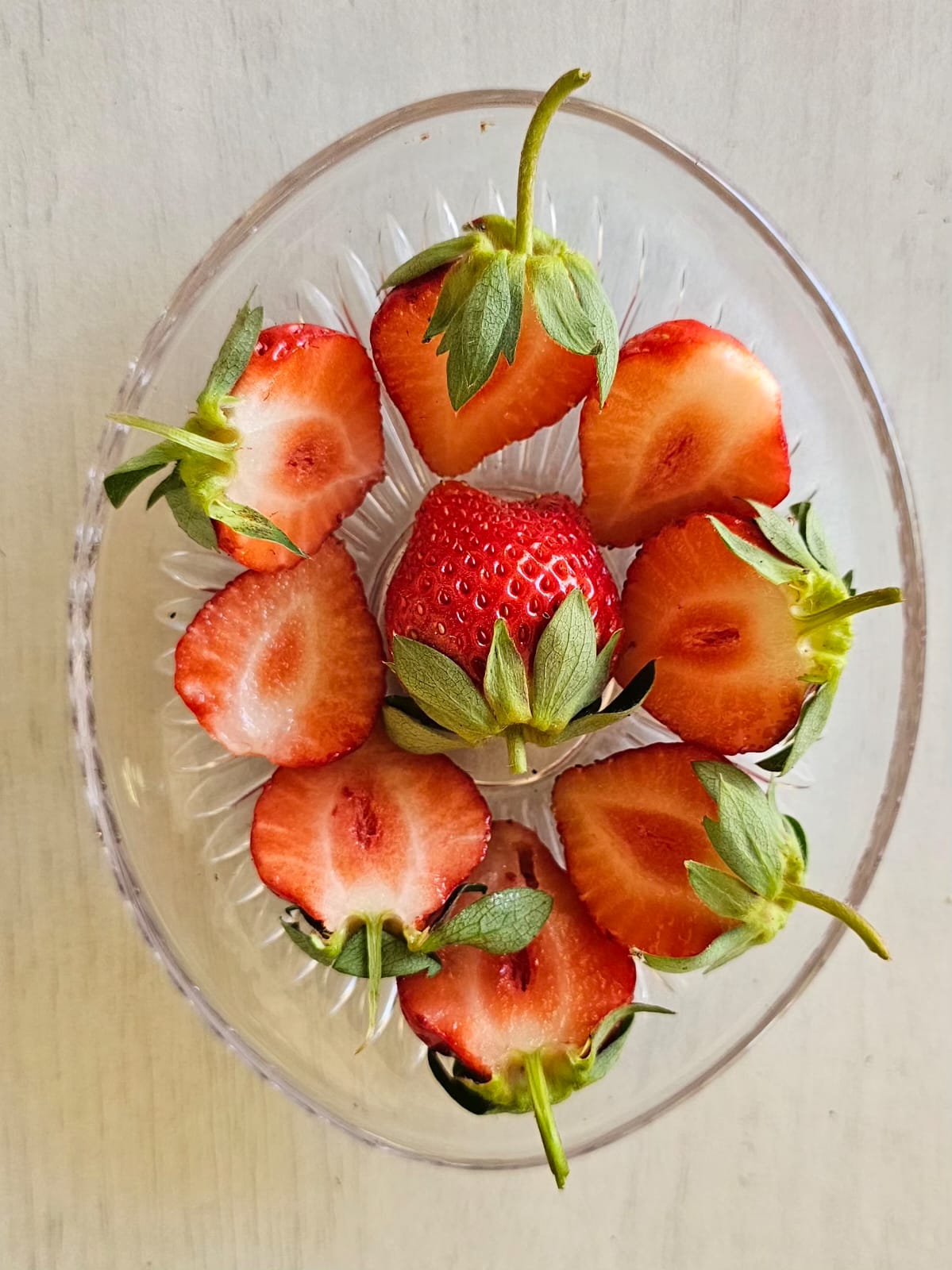A glass dish with one whole strawberry at the centre surrounded by six sliced halves showing the deep red cross-sections